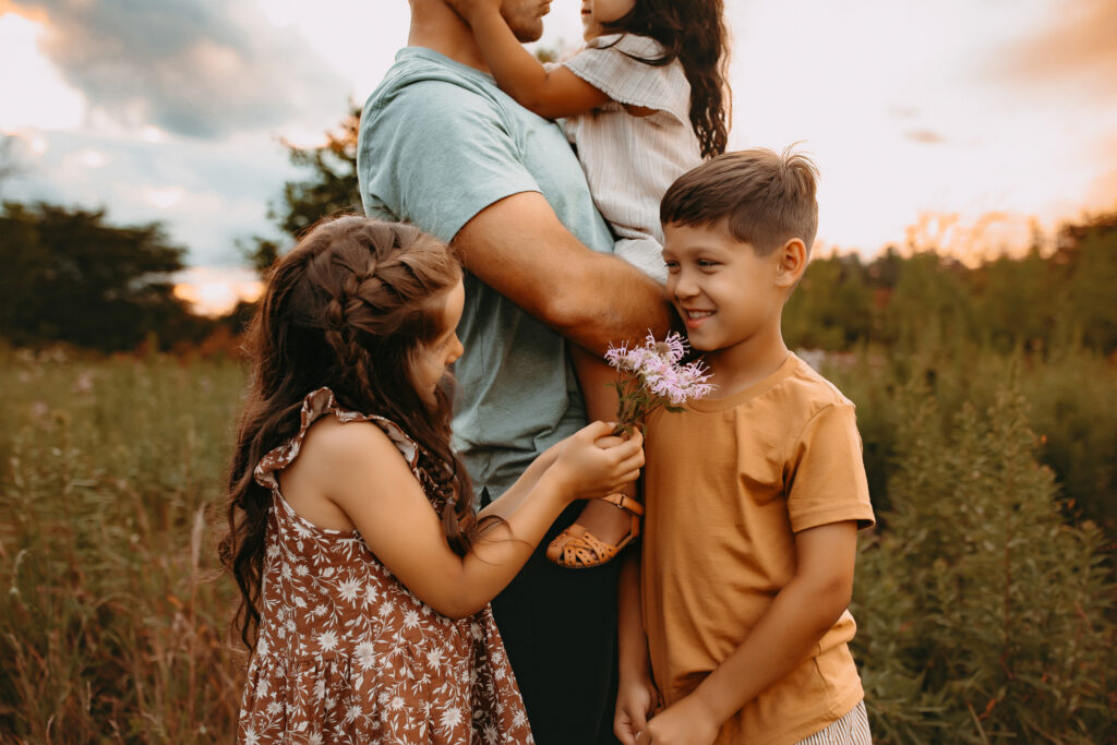 Little sister offering pretty purple wild flowers to her older brother and their father holds their little sister behind them. 