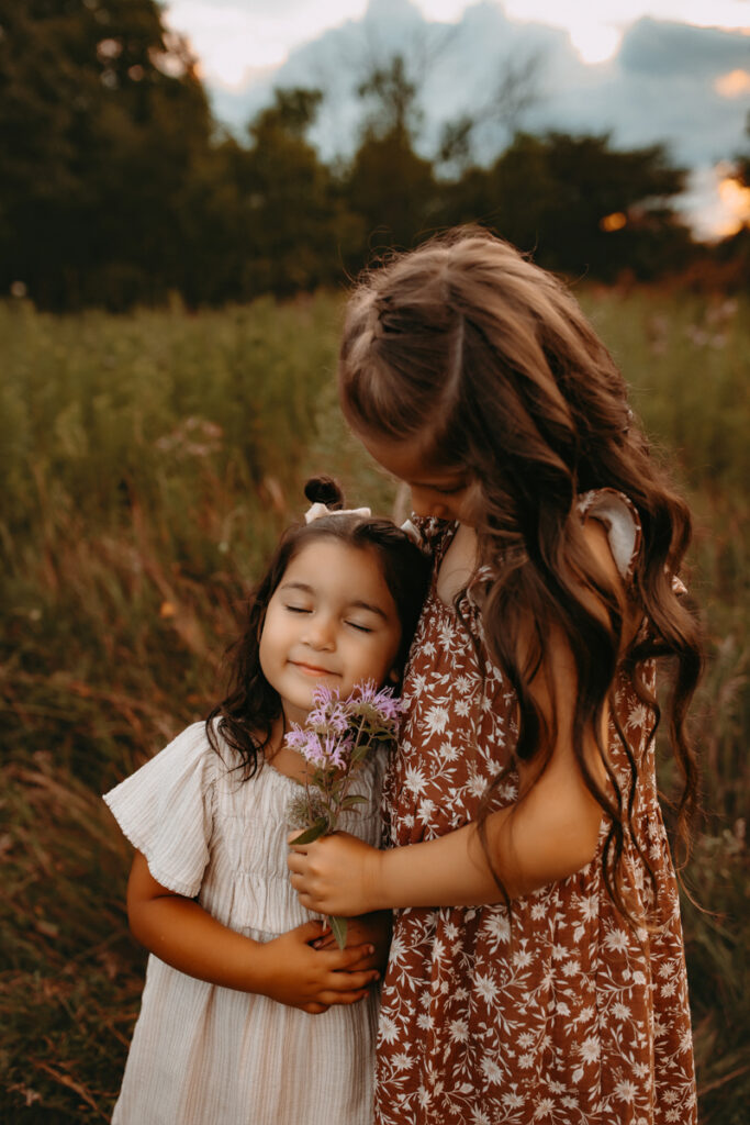 Older sister holding her little sister and handing her purple wildflowers. 